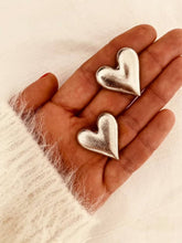 Load image into Gallery viewer, Pair of oversized silver heart statement earrings with a brushed metallic finish, photographed on hand.