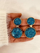 Load image into Gallery viewer, NYOKO blue resin statement earrings displayed in a hand, highlighting the sparkling resin texture and gold-tone finish.
